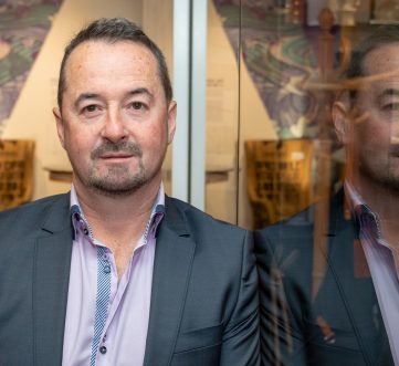 Gerry Hughes standing next to a glass wall in which you can see his reflection