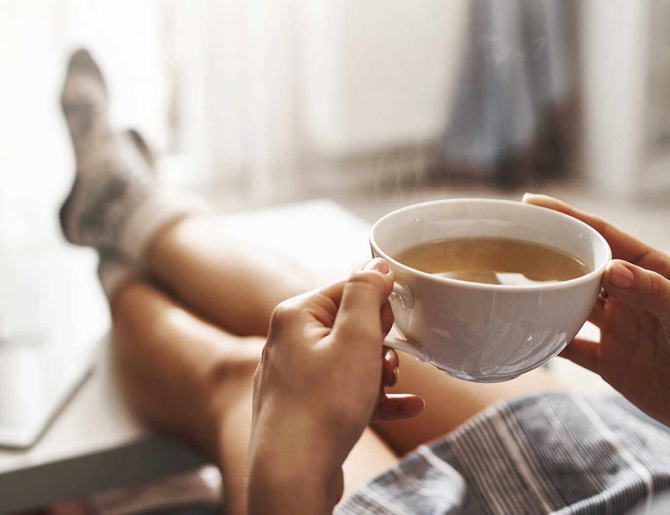 woman holding a cup of tea with her legs stretched out on the table in front of her