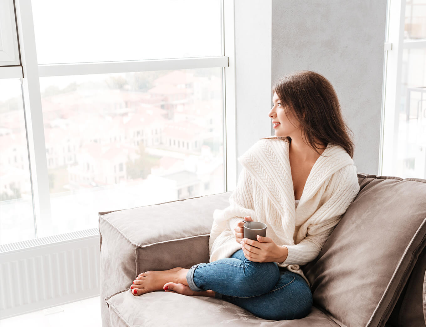 woman sitting curled up on a couch, drinking a cup of tea and looking out the window