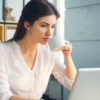 woman taking a sip of tea while working at a laptop