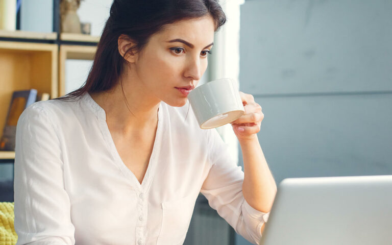 woman taking a sip of tea while working at a laptop