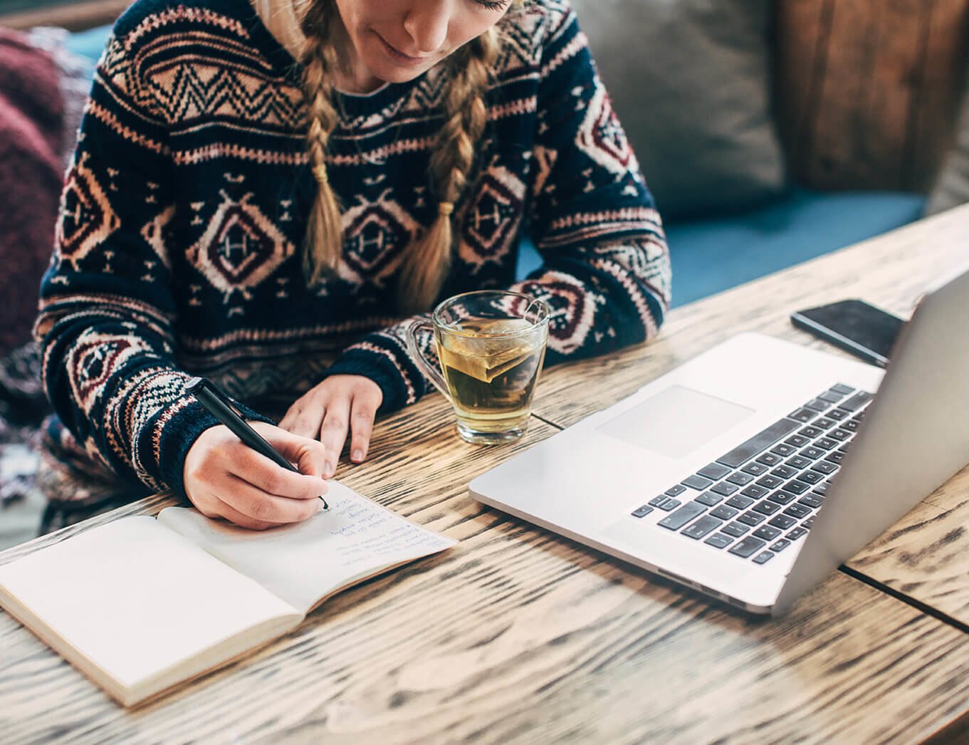 woman writing in a copy book with a laptop and a glass of tea in front of her