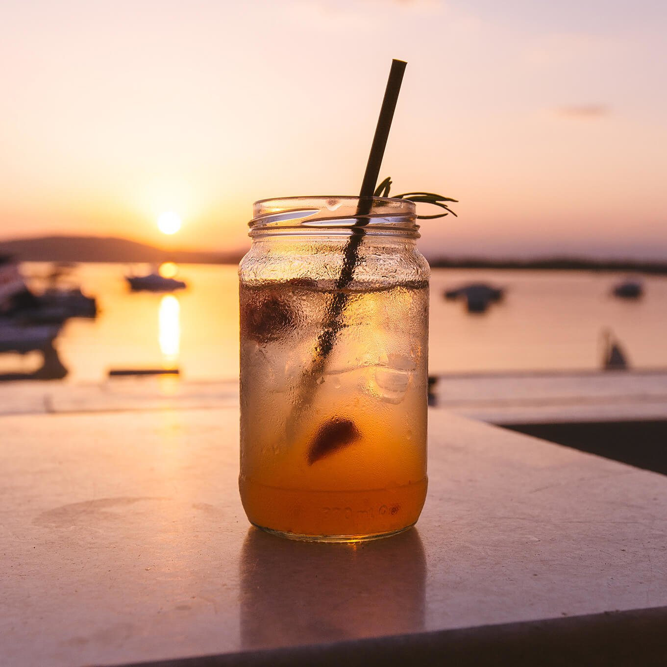 clear jar with iced tea and a straw, on a table with a beautiful sunset over the sea in the background