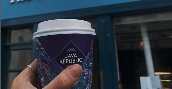 Hand holding a Java Republic coffee cup in front of a shop front ' The Daily Grind'