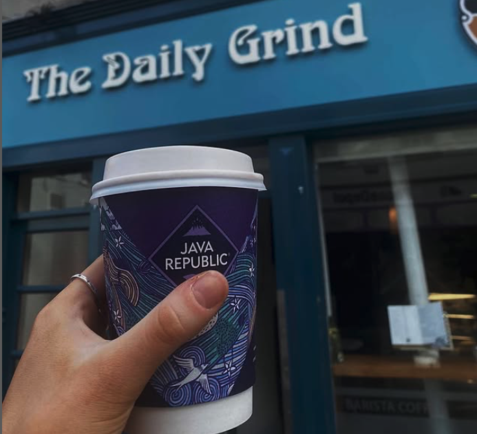 Hand holding a Java Republic coffee cup in front of a shop front ' The Daily Grind'